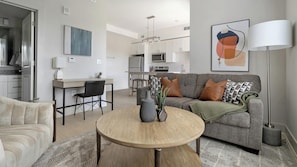 Ein modernes Wohnzimmer mit grauem Sofa, einem runden Holztisch und einem Essbereich mit weißem Tisch und Stühlen.