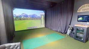 Ein Raum mit einem großen Bildschirm, der einen Golfplatz zeigt, einer Golfbag und einem Monitor auf einem Schreibtisch.