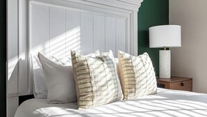 A bed with white bedding, striped pillows, a white nightstand, a white lamp, and a green wall. - Landing | Stylish 2BD, Game Room, Gym (Charlotte)