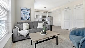 Een moderne woonkamer met een grijze bank, een salontafel en een blauwe fauteuil. Achtergrond toont een keuken met witte kastjes en een magnetron.