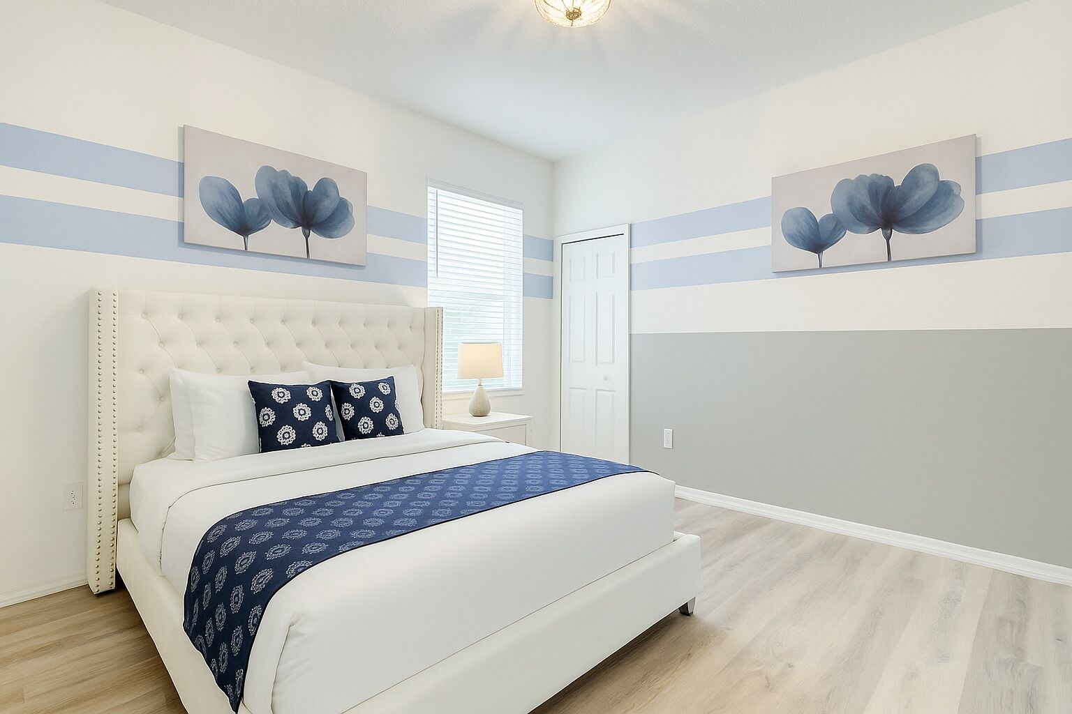 Quarto com cabeceira estofada, cama com almofadas estampadas, mesa de cabeceira com abajur e dois quadros de flores pendurados na parede.