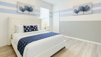 Quarto com cabeceira estofada, cama com almofadas estampadas, mesa de cabeceira com abajur e dois quadros de flores pendurados na parede.