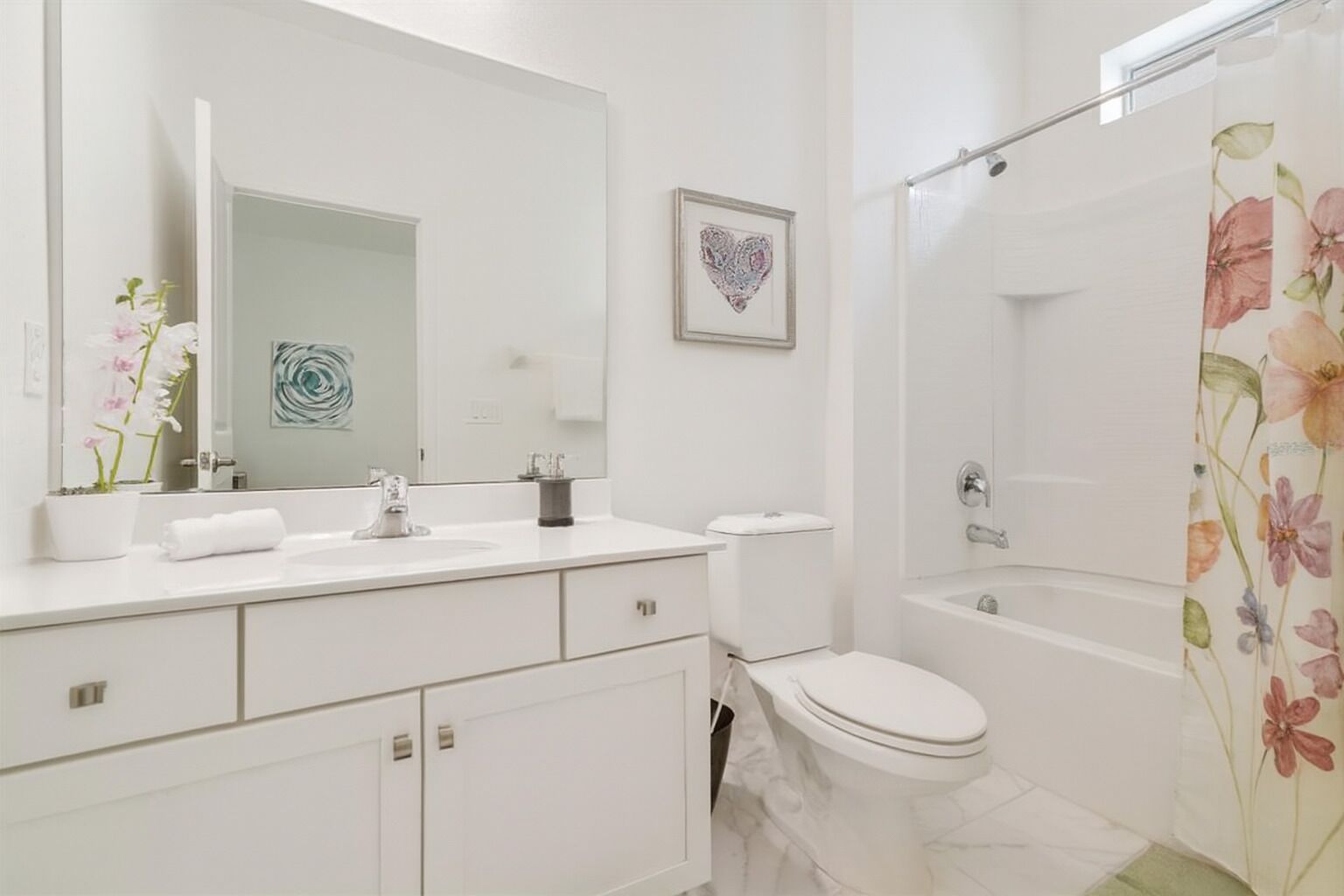 A modern bathroom with a white sink, toilet, and bathtub. There is a heart-shaped artwork on the wall and a floral shower curtain.