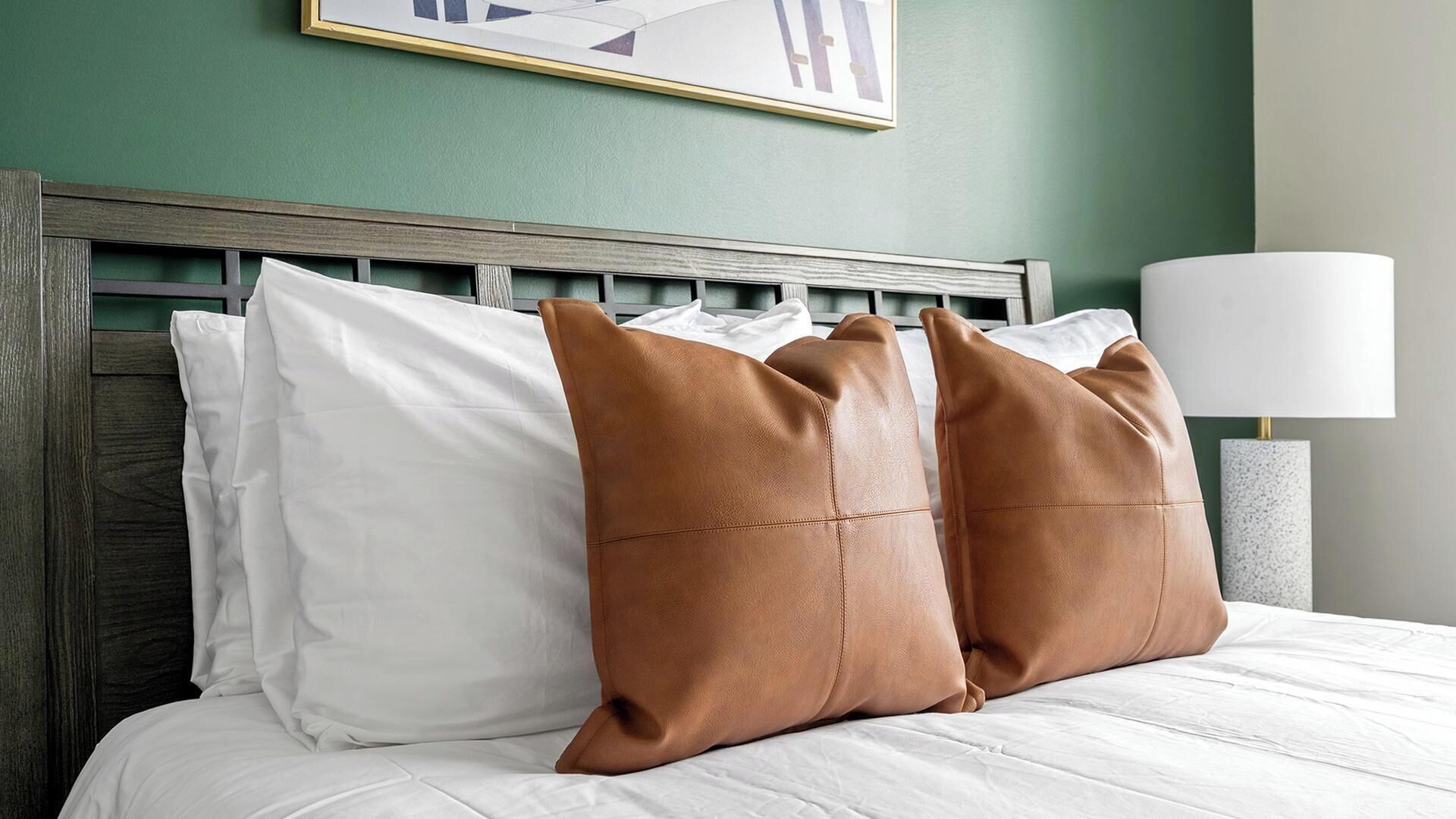 A bed with white linens and two brown pillows, a wooden headboard, and a wall-mounted lamp.