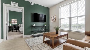 A modern living room with a TV, sofa, coffee table, and a view of the city through large windows. - Landing | Amazing 1BD, Gym, Game Room (Charlotte)