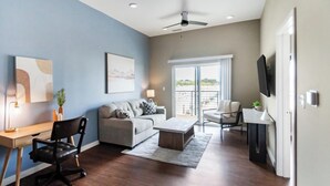 A modern living room with a sofa, coffee table, and TV, a balcony with a view, and a desk with a chair. - Landing | Beautiful 1BD, Rooftop Lounge, Gym (Springfield)