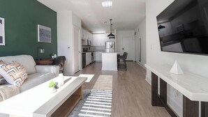 Un salón moderno con sofá, mesa de centro y televisión. Se ve una cocina en el fondo, con armarios y electrodomésticos blancos.