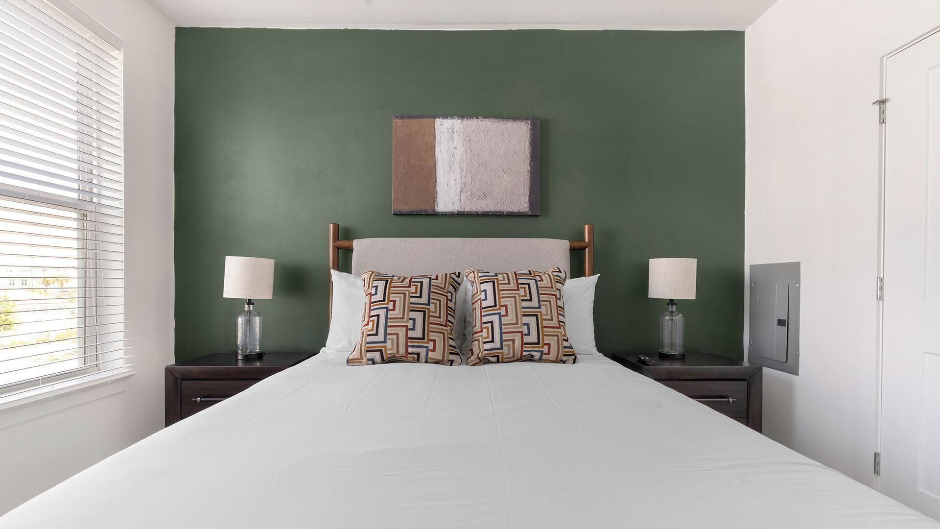 A bedroom with a bed, two nightstands, a lamp on each, a painting above the bed, and a window with blinds.