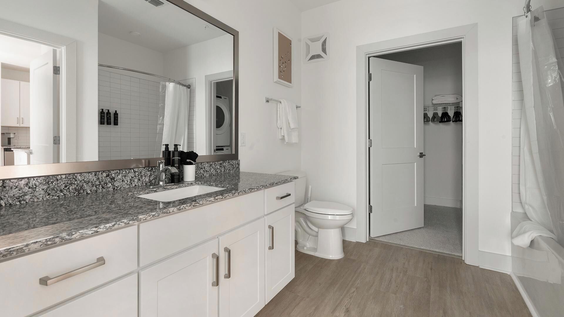 Un baño moderno con encimera de granito, muebles blancos, un espejo grande y cortina de ducha.