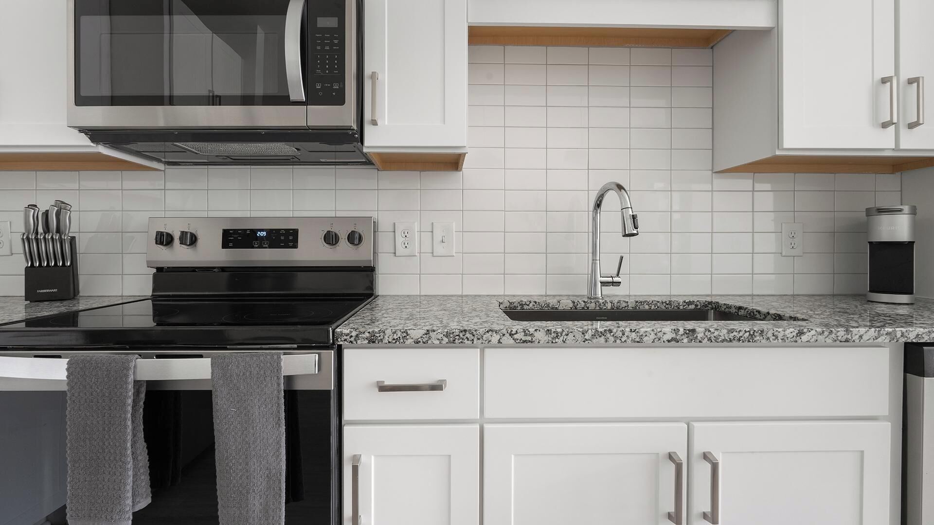 Eine moderne Küche mit weißen Schränken, einer Edelstahl-Mikrowelle, einem Backofen, einer Granit-Arbeitsplatte, einer Spüle und einer Kaffeemaschine.