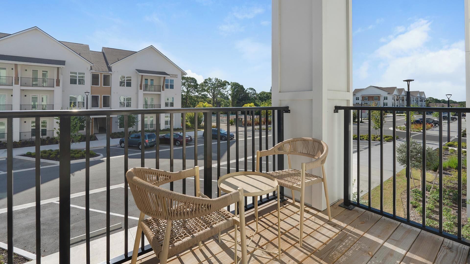 En balkon med to fletstole, udsigt til en parkeringsplads og bygninger i baggrunden.