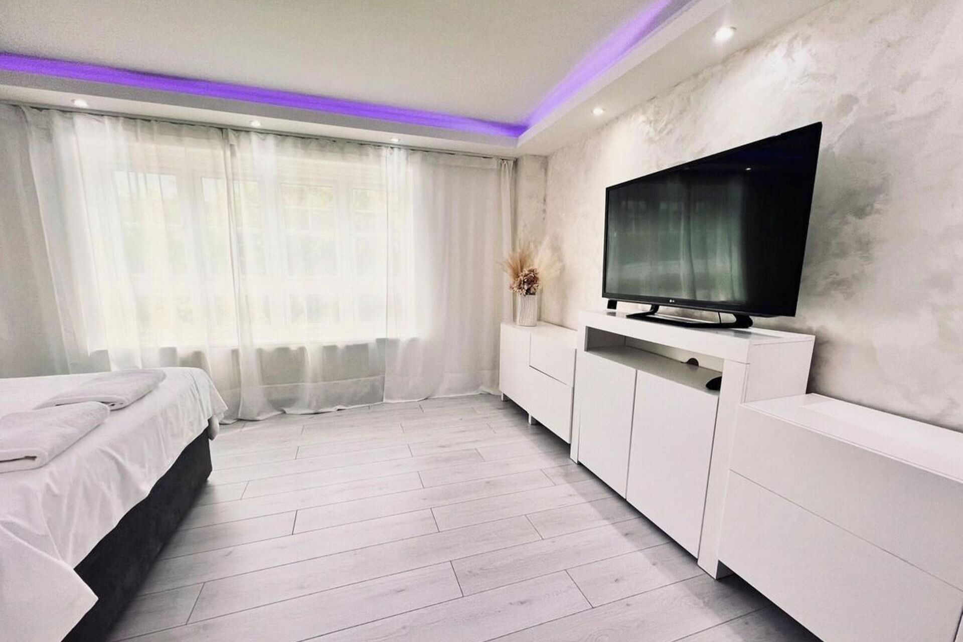 Une chambre moderne avec un lit, une télévision, une armoire blanche et une grande fenêtre ornée de rideaux légers.