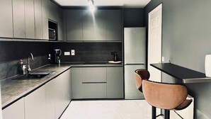 Una cocina moderna con armarios grises, encimera negra, un lavamanos, cafetera y refrigerador. Hay dos taburetes de bar con asiento de cuero marrón.