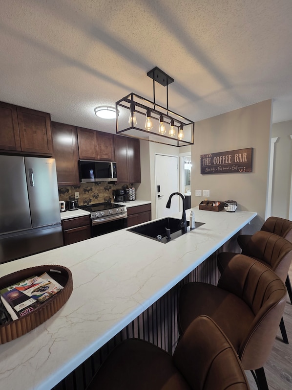 A modern kitchen with a central island, dark wooden cabinets, a stainless steel refrigerator, a microwave, and a coffee bar sign.