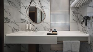 Une salle de bain avec un mur en marbre, un grand miroir, un lavabo blanc, un porte-serviettes et un sèche-cheveux.