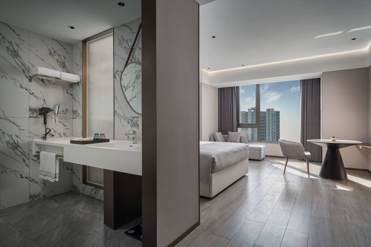 A modern hotel room with a marble bathroom, a bed, a dining area, and a view of high-rise buildings.