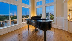 A grand piano in a spacious room with large windows offering a view of the ocean. - Oceanfront Estate | Sleeps 40 | Weddings & Retreats | 18BR | OBX (Corolla)