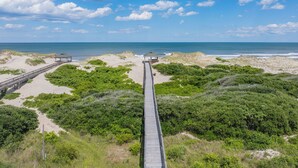 A wooden boardwalk leading to beach huts, surrounded by dense greenery and sandy dunes. - Oceanfront Estate | Sleeps 40 | Weddings & Retreats | 18BR | OBX (Corolla)
