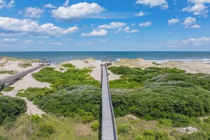 Ein hölzerner Steg führt zu Strandhäusern, umgeben von dichter Vegetation und Sanddünen.