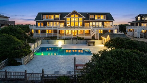 A large, multi-story house with a swimming pool, multiple balconies, and a well-lit exterior. - Oceanfront Estate | Sleeps 40 | Weddings & Retreats | 18BR | OBX (Corolla)