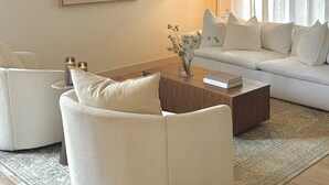 Ein modernes Wohnzimmer mit einem runden weißen Sofa, einem Holztisch und einem weißen Sessel. An der Wand hängt ein Gemälde und auf dem Tisch steht eine Lampe.