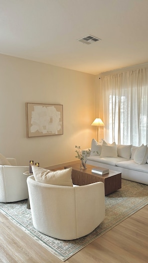 Ein modernes Wohnzimmer mit einem runden weißen Sofa, einem Holztisch und einem weißen Sessel. An der Wand hängt ein Gemälde und auf dem Tisch steht eine Lampe.