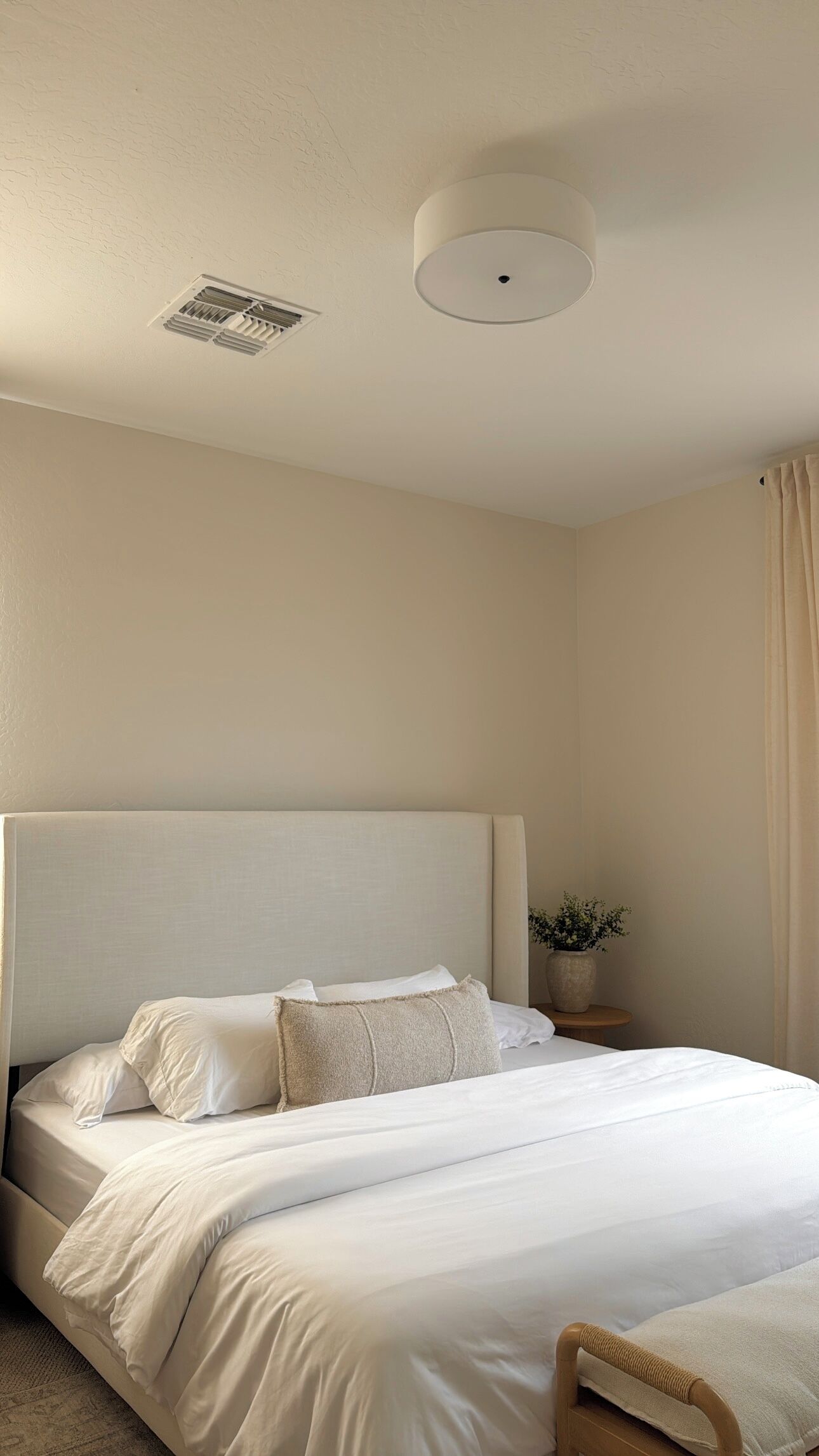 A hotel room with a large bed, a bedside table with a vase, and a ceiling vent.