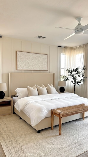 Ein Schlafzimmer mit einem Bett, Nachttischen, einer Bank, einem Deckenventilator, einem Fenster mit Vorhängen und einem Wandbild.