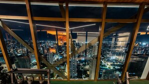 Academia com um banco, kettlebells e uma estrutura para agachamento, com vista para o horizonte urbano à noite.