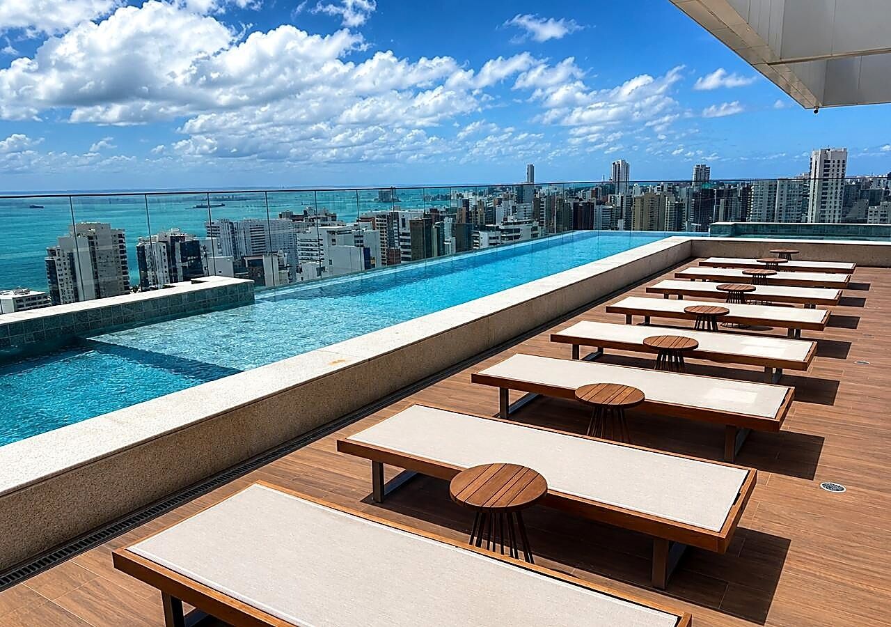 A rooftop pool with lounge chairs and tables, overlooking a city skyline and the ocean.