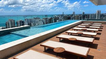 A rooftop pool with lounge chairs and tables, overlooking a city skyline and the ocean.