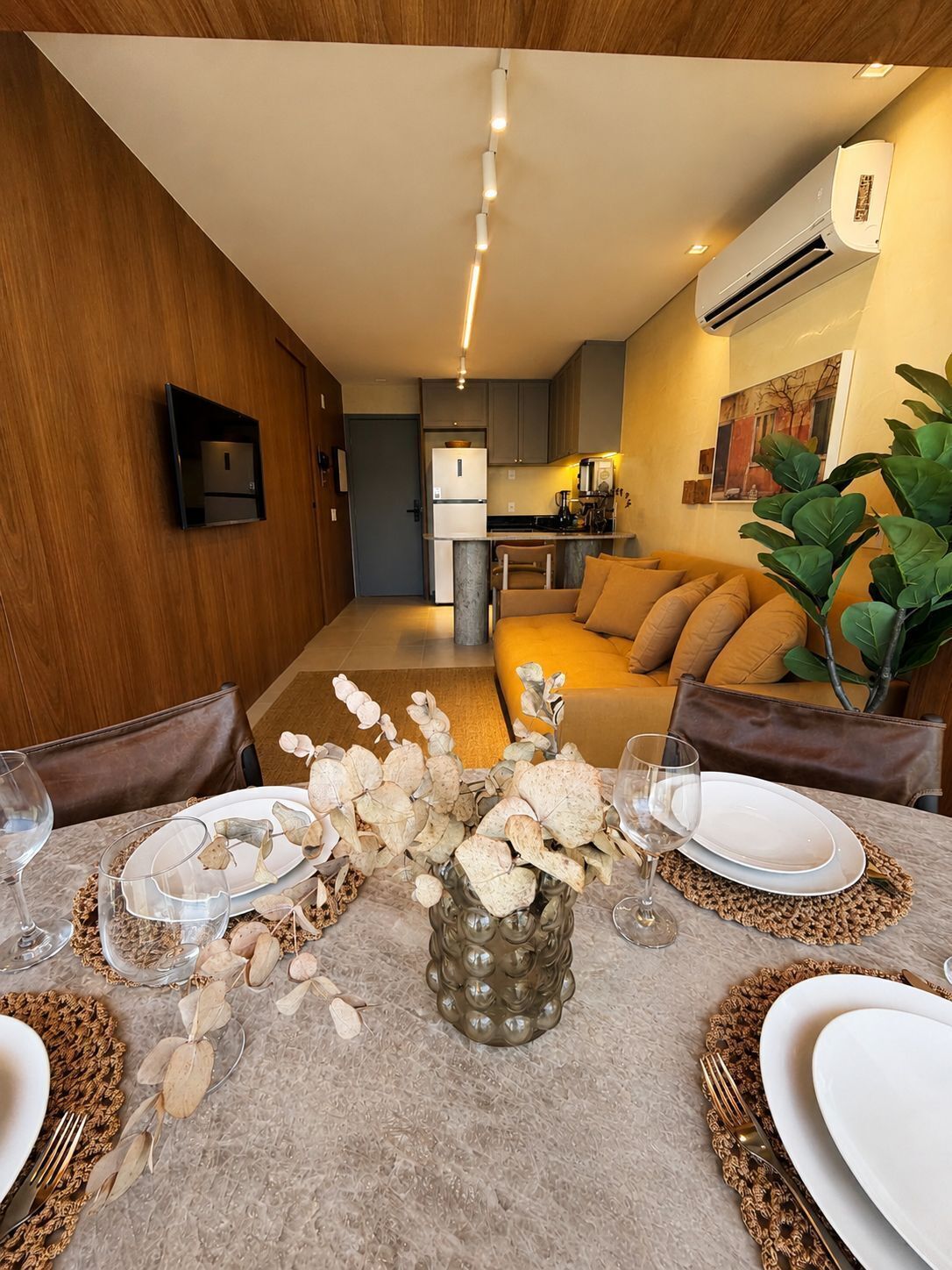 Une cuisine moderne dotée d’un four encastré, un coin repas avec une table préparée pour deux personnes, et un salon avec un canapé et une plante.
