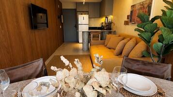 Une cuisine moderne dotée d’un four encastré, un coin repas avec une table préparée pour deux personnes, et un salon avec un canapé et une plante.