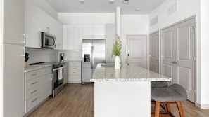 A modern kitchen with white cabinetry, stainless steel appliances, a granite countertop island, and a wooden stool. - Landing | Spectacular 1BD, Clubhouse, Gym (Jacksonville)