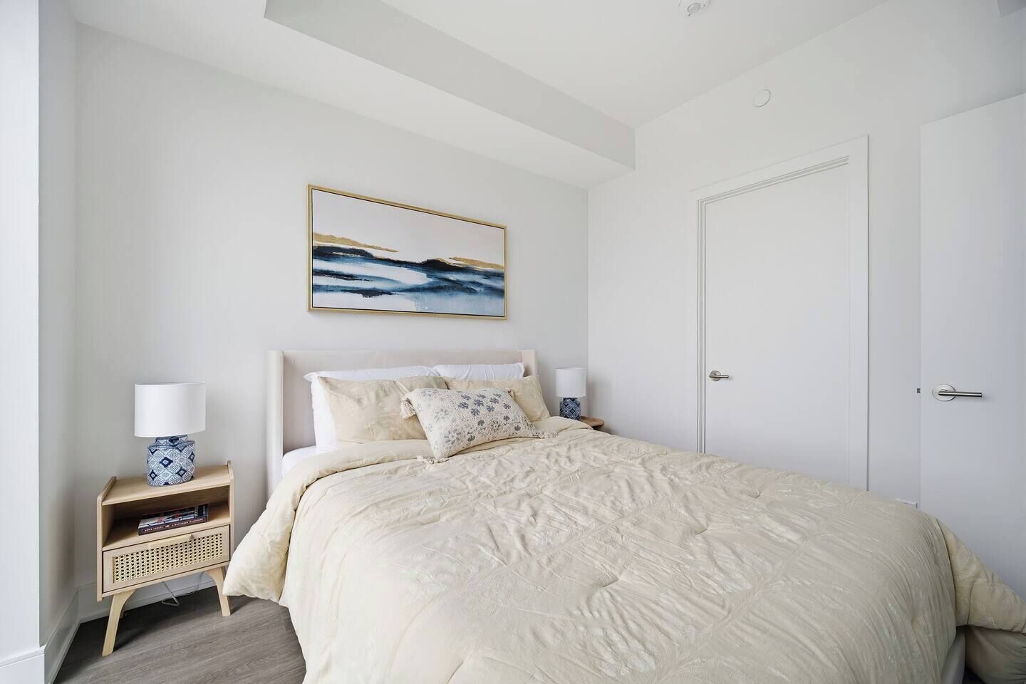 A bedroom with a bed, a nightstand, a lamp, a framed picture on the wall, and a door.