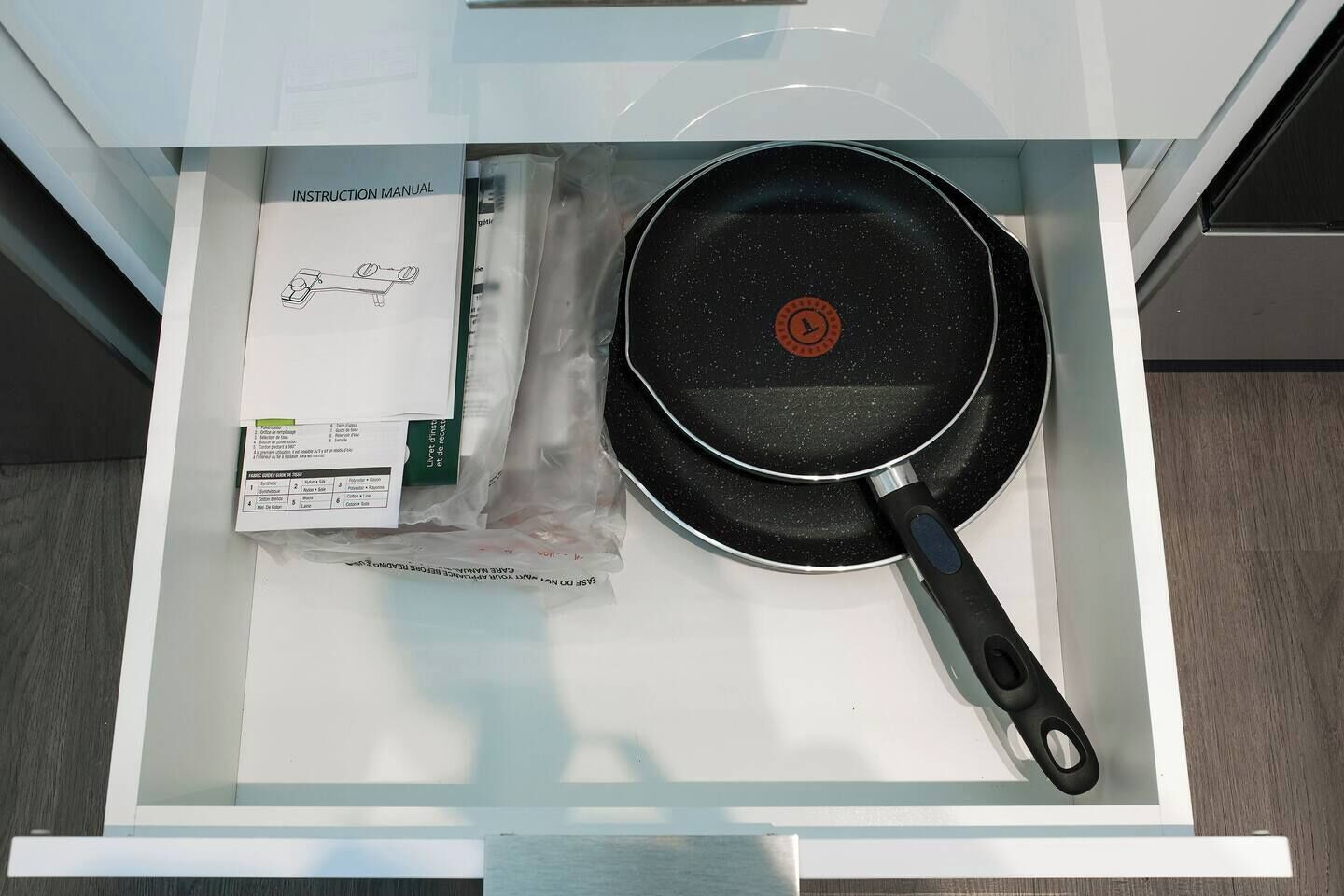 A kitchen drawer containing a frying pan, an instruction manual, and a plastic bag.