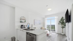 Una cocina moderna con lavavajillas, pileta y cafetera. Hay un comedor con mesa y sillas, y un amplio ventanal con vistas a la ciudad.