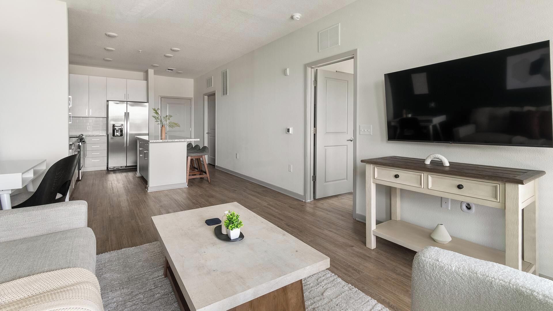 A modern living room with a flat-screen TV, a dining area with a kitchen in the background, and a small bar area.