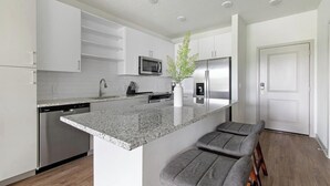 Una cocina moderna con armarios blancos, encimera de granito, microondas integrado, refrigerador de acero inoxidable y una isla central con taburetes de bar.