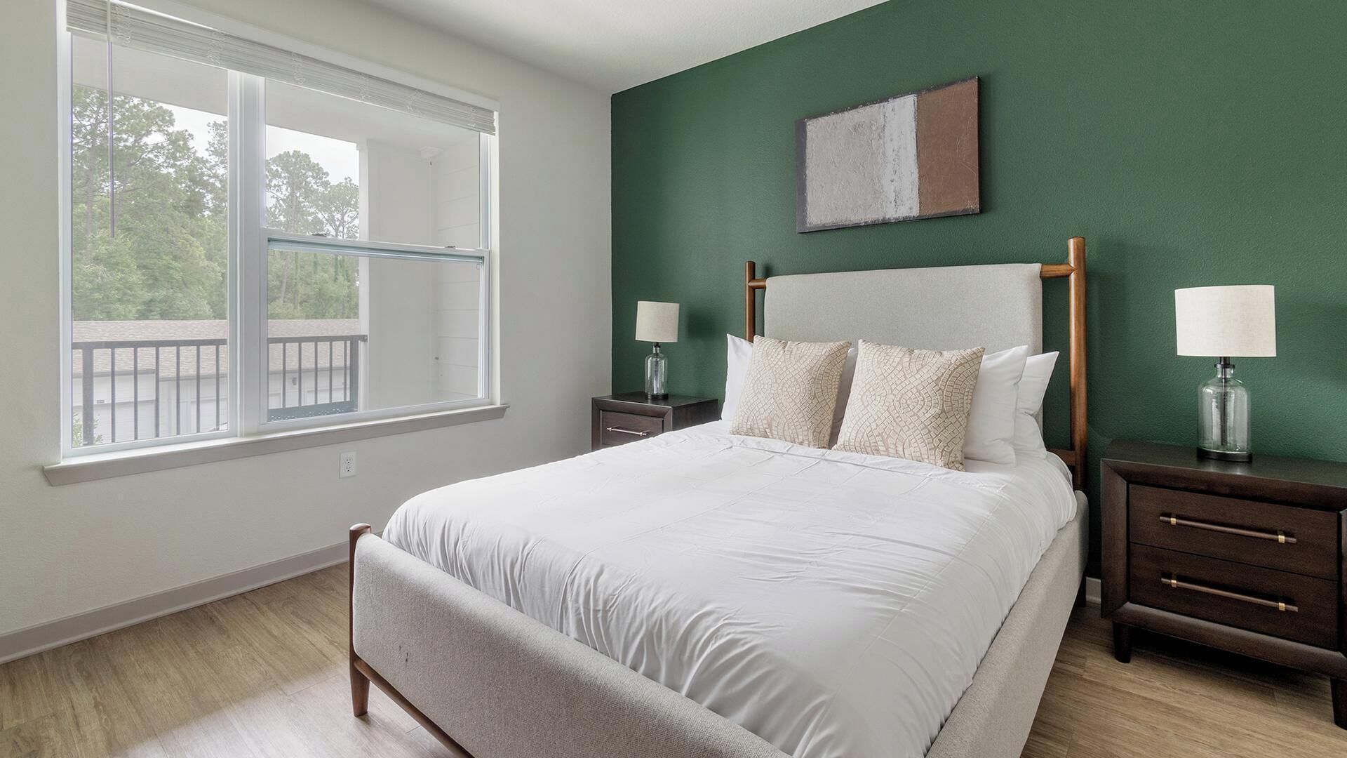 A bedroom with a large bed, two bedside tables with lamps, a window with a view, and a green accent wall.
