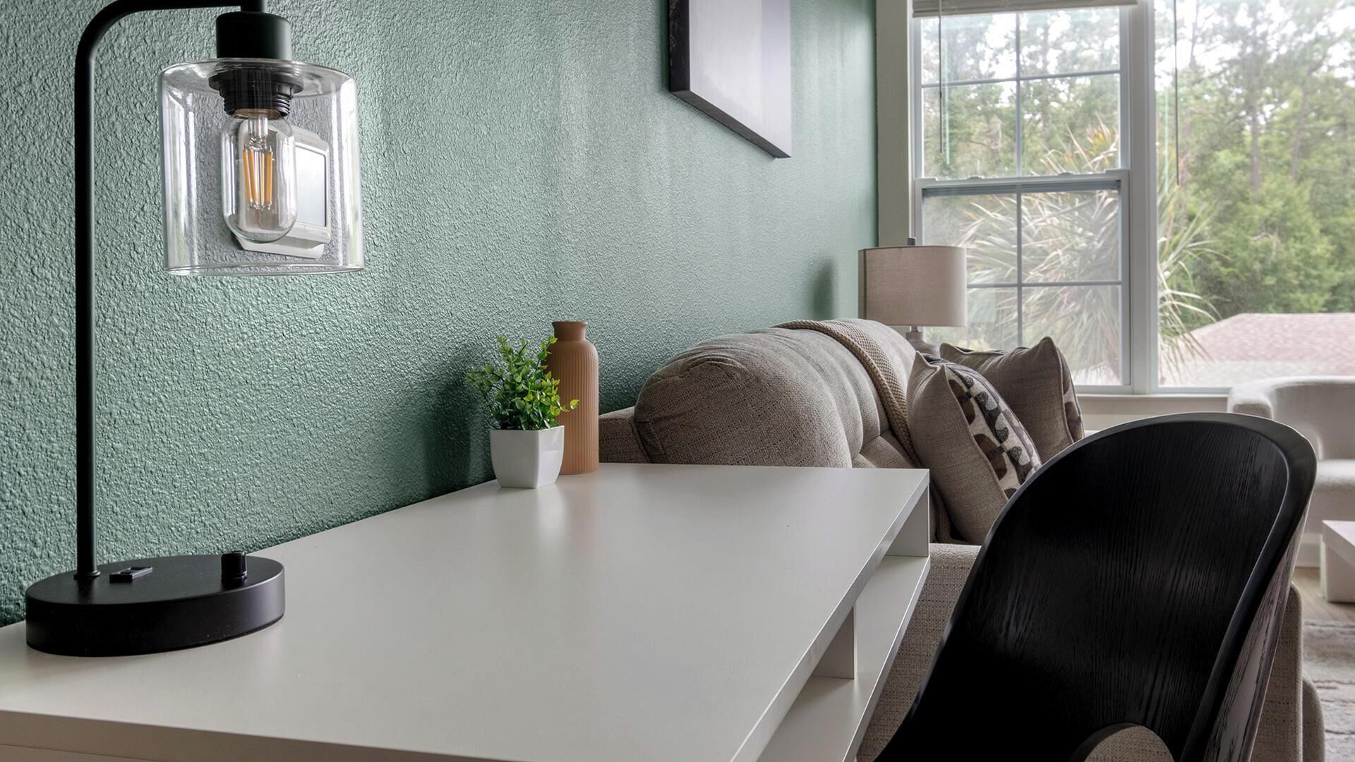 Ein modernes Zimmer mit Schreibtisch, Lampe, Blumentopf und einer Couch mit Kissen.