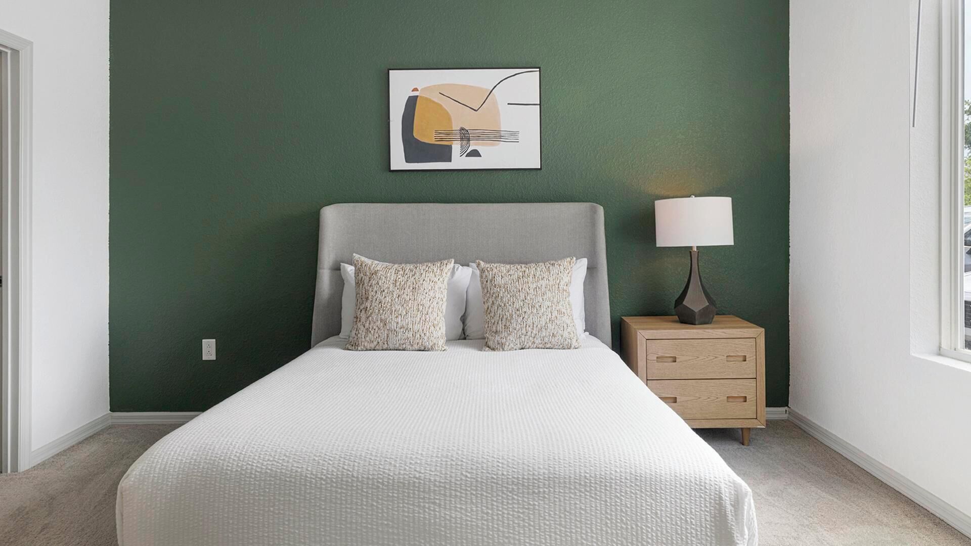 A bedroom with a bed, two pillows, a nightstand with a lamp, and a wall art piece.