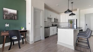 Una cocina moderna con armarios blancos, electrodomésticos de acero inoxidable, una zona de comedor con escritorio y sillas, y una isla grande con taburetes de bar.