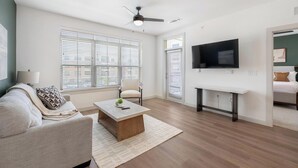 A modern living room with a sofa, coffee table, TV, and large windows with blinds. - Landing | Stunning 2BD, Gym, Clubhouse (Celina)