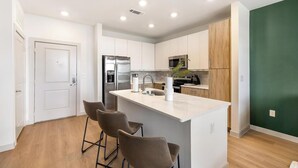 A modern kitchen with white cabinets, a stainless steel refrigerator, a built-in oven, and a bar area with stools. - Landing | Amazing 1BD, Courtyard, Clubhouse (Celina)