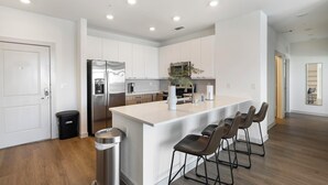 A modern kitchen with a stainless steel refrigerator, a central island with bar stools, and a trash can. - Landing | Amazing 3BD, Clubhouse, Gym (Celina)