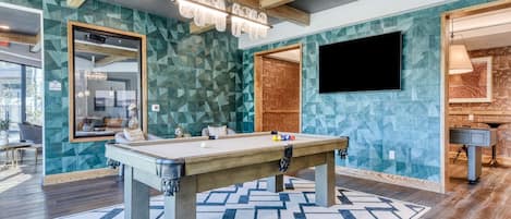 A pool table in a room with geometric patterned walls, a mounted TV, and a visible door leading to another area.