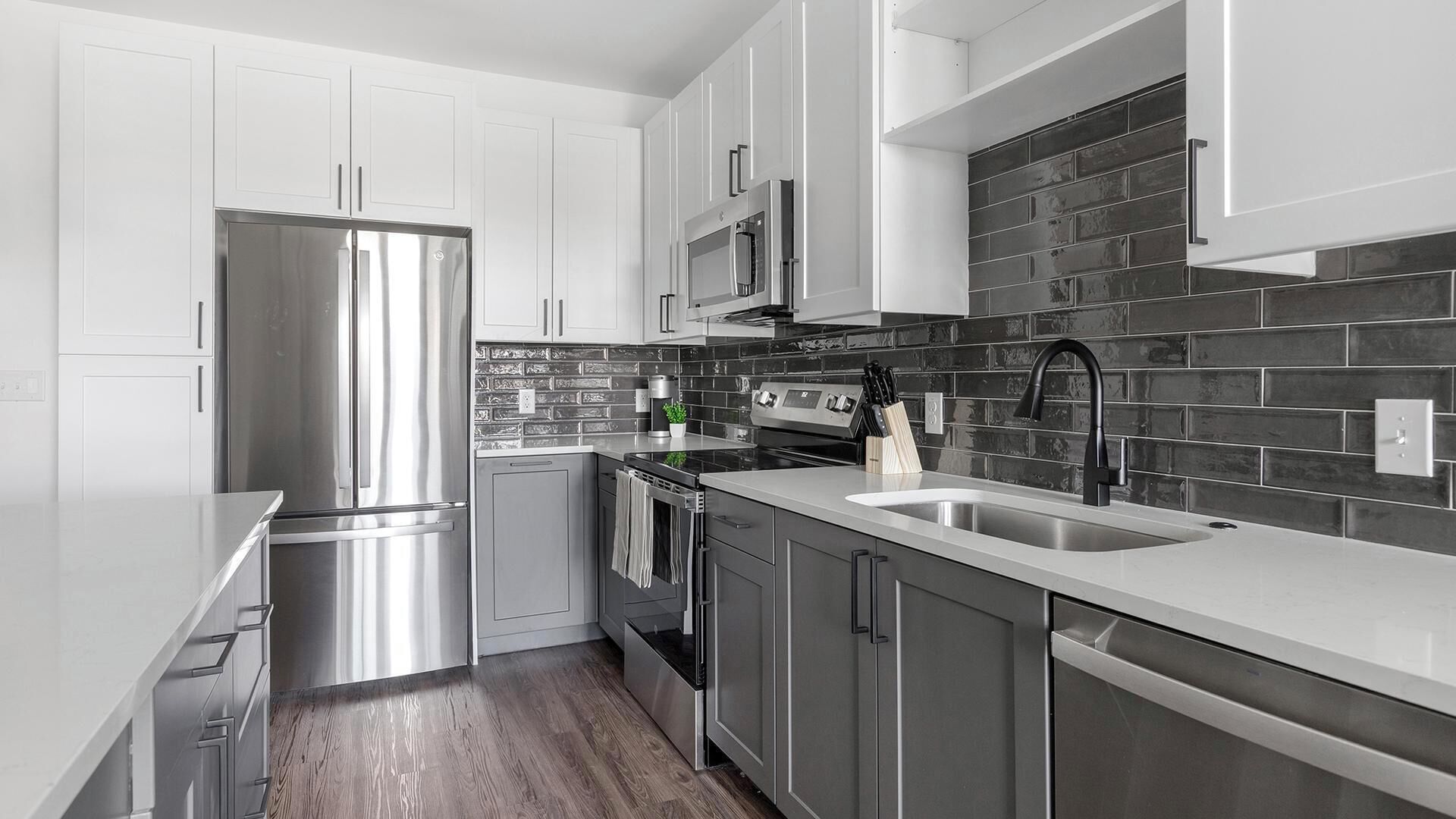 A modern kitchen with stainless steel appliances, a black tiled backsplash, and wooden flooring.
