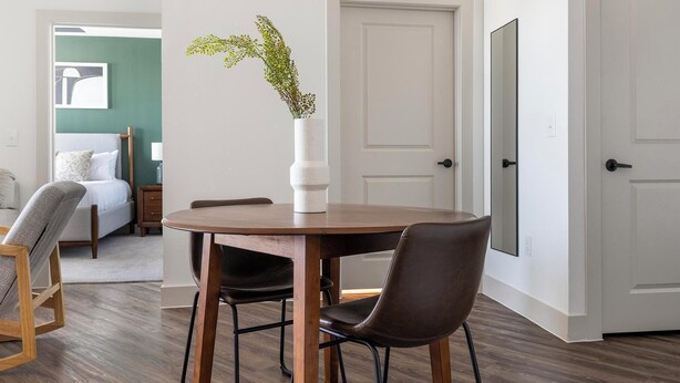 A dining area with a round table, two chairs, a vase with a plant, a bed with a pillow, a dresser, and a door. - Landing | Incredible 1BD, Yoga Studio (San Antonio)