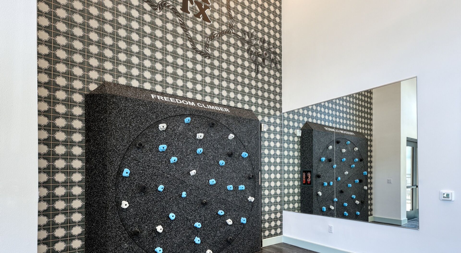 Un mur d’escalade à surface noire et à prises bleues, portant l’inscription « FREEDOM CLIMBER », dans une pièce aux murs décorés de motifs.